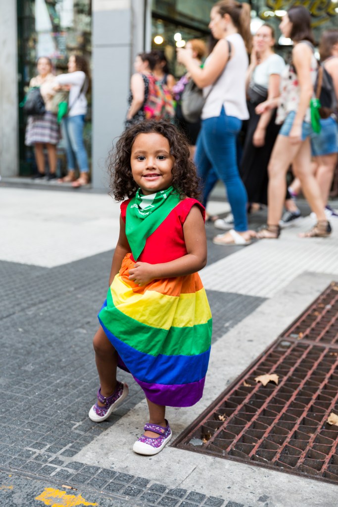 Une petite fille un foulard vert autour du coup et une robe aux couleurs de l'arc-en-ciel