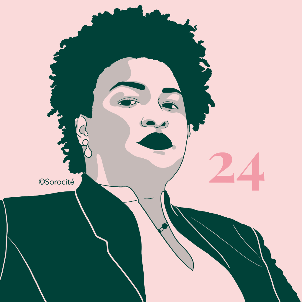 Stacey Abrams en format portrait vue légèrement depuis le bas, perles aux oreilles, haut clair et veste foncée, ses cheveux naturels mi-longs. 